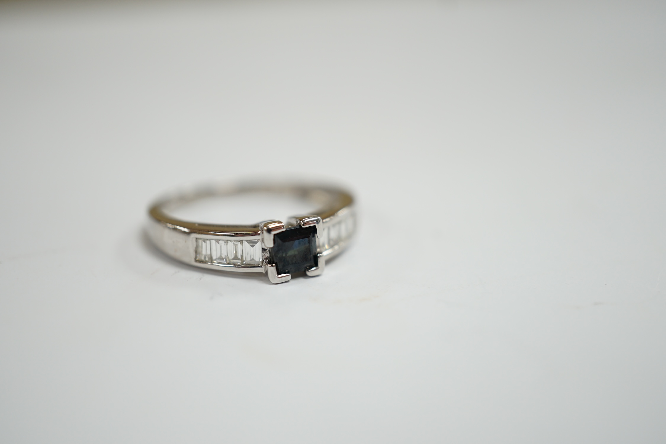 A modern 18ct white gold and single stone square cut sapphire set ring, with baguette cut diamond set shoulders, size N, gross weight 2.9 grams.